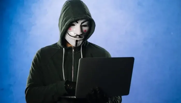 Imagem de homem de máscara e capuz com notebook na mão em um fundo azul ilustrando o que é peculato.